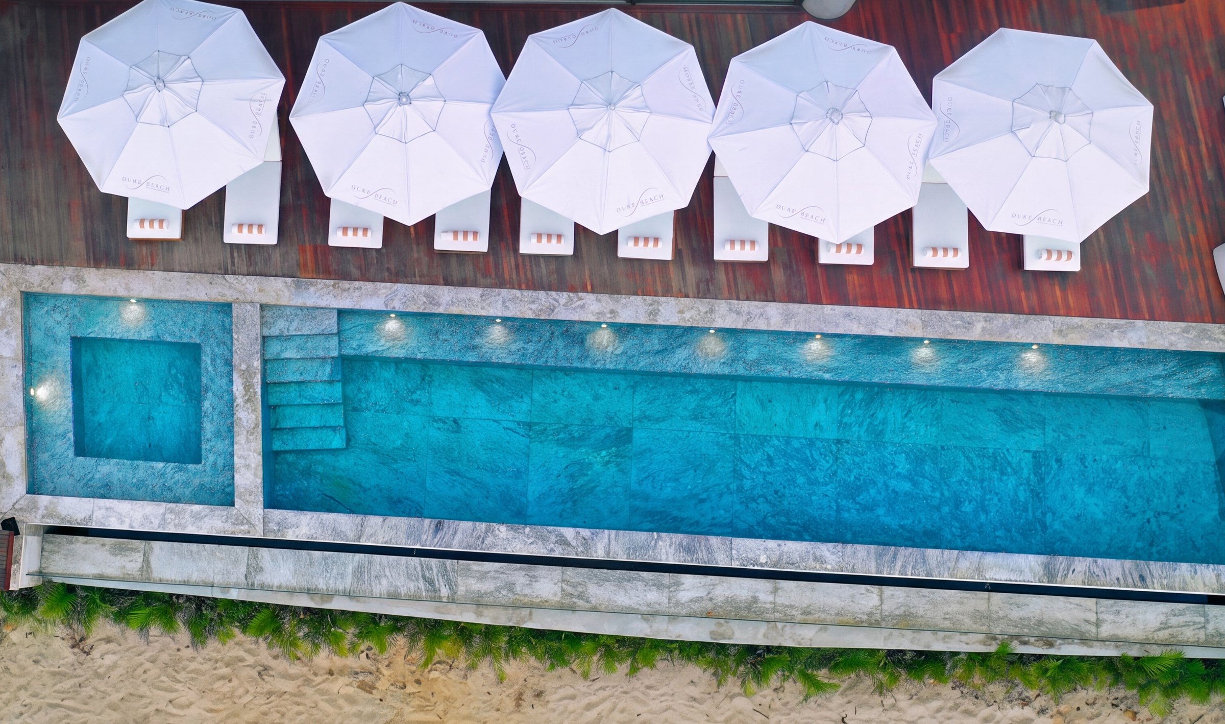 A piscina de borda infinita do Duke Beach vista de cima: cinco guarda-sóis brancos com a marca do hotel, as espreguiçadeiras alinhadas com toalha enrolada e, abaixo, a faixa de areia da praia de Maresias.