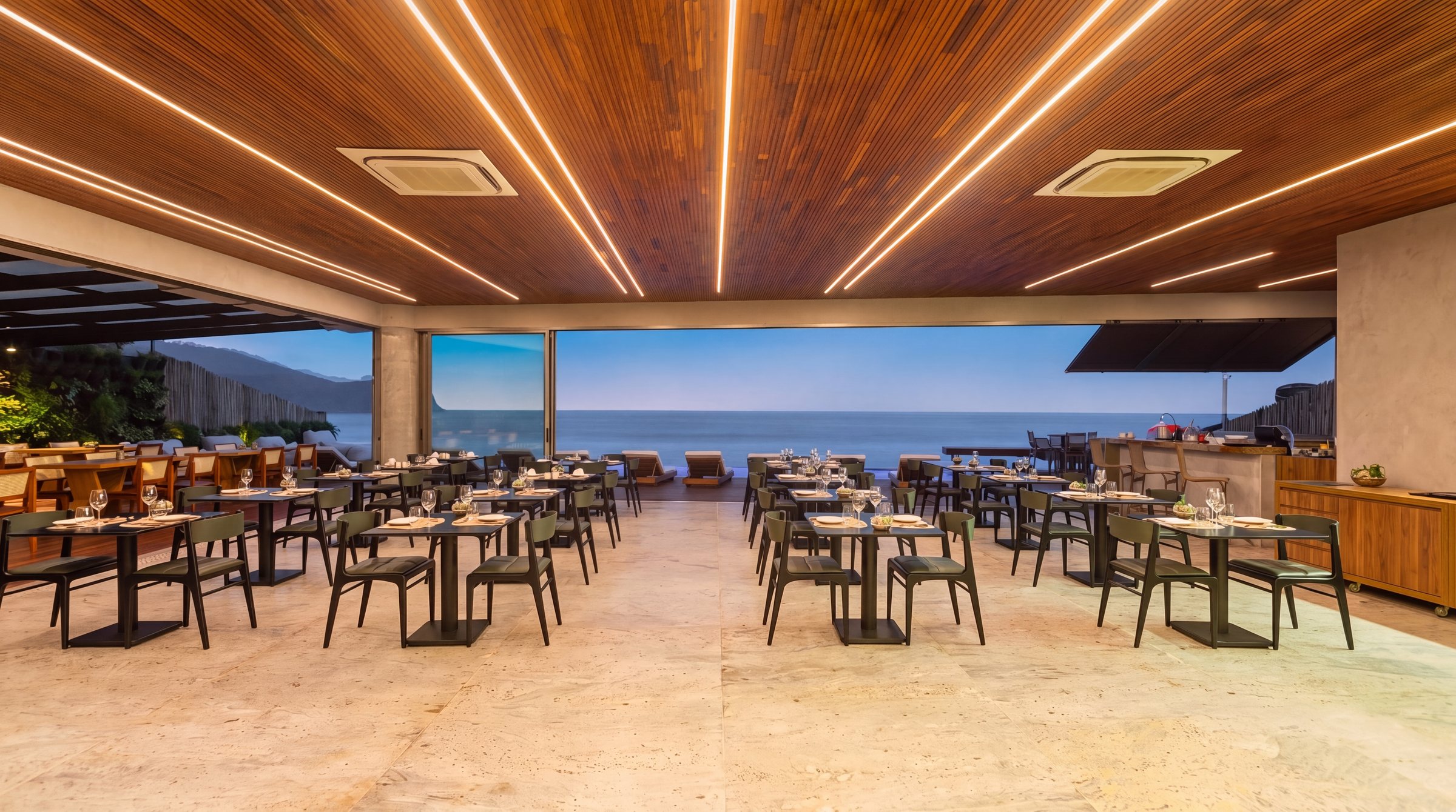 O restaurante do Duke Beach com as mesas postas, o forro de madeira com linhas de luz e, ao fundo, as janelas abertas para o mar de Maresias.